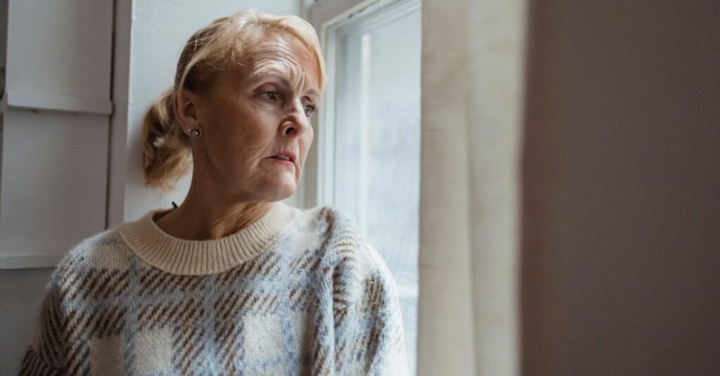 Contemplative senior female wearing warm sweater sitting on windowsill and looking away in sad thoughts