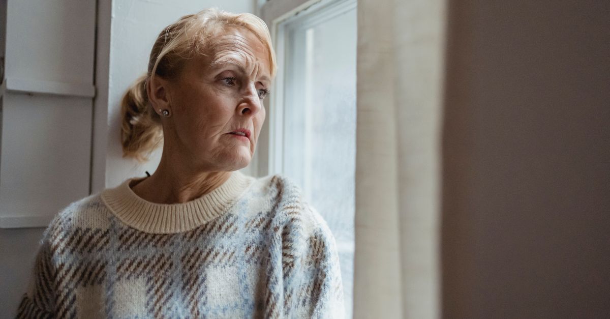 Contemplative senior female wearing warm sweater sitting on windowsill and looking away in sad thoughts