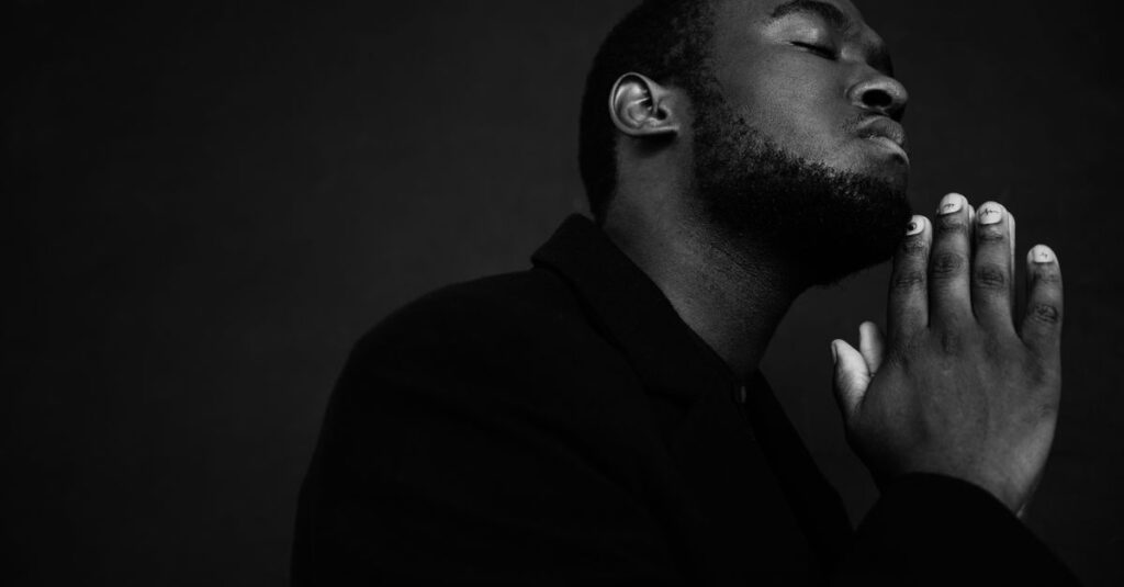 Monochrome handsome unshaven African American male in black suit praying with hands clasped under chin