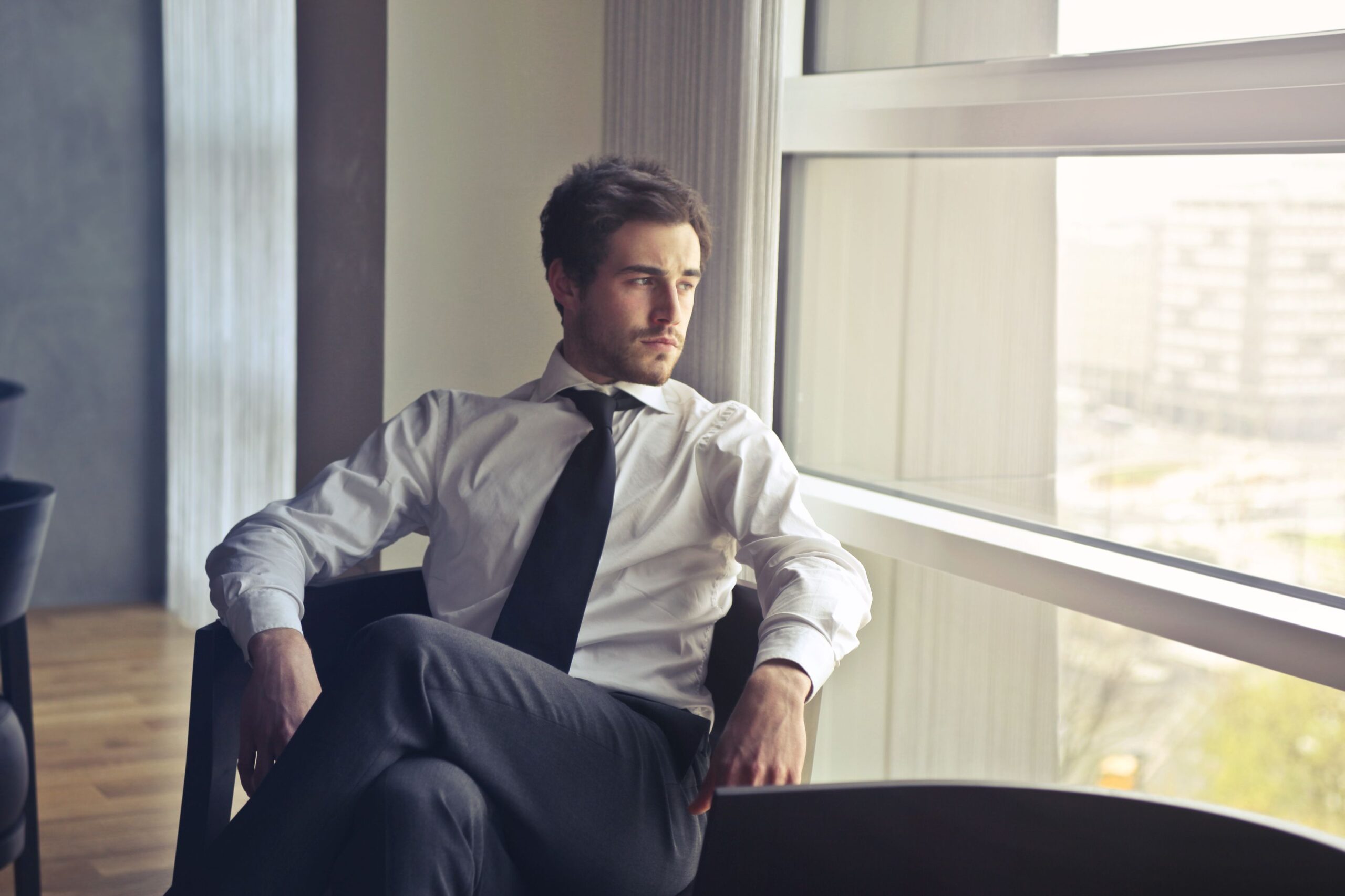 Young businessman in formal attire sitting and looking out of modern office window, contemplating.